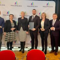 Zdjęcie przedstawiające od prawej Kierownik Magdalena Doniec, Dyrektor Roman Wcisło, Wiceprezes Blisko Krakowa Agata Kowalska, Prezes Blisko Krakowa Michał Kędzierski, Członek Zarządu Województwa Małopolskiego Iwona Gibas, Skarbnik Blisko Krakowa Małgorzata Tomczyk, Wiceprezes Blisko Krakowa Stanisław Kozioł razem na tle ścianki województwa małopolskiego